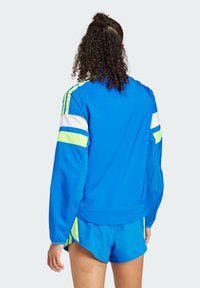 Chaqueta deportiva azul con franjas amarillas neón y blancas en los hombros. Tejido ligero con un diseño deportivo, combinada con pantalones cortos a juego.