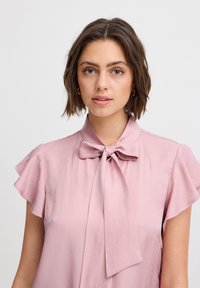 Blusa rosa chiaro con maniche corte a campana e colletto con fiocco annodato. Tessuto liscio, leggermente lucido, con una vestibilità morbida. Cuciture e dettagli semplici.