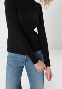 Schwarzes Langarmoberteil aus geripptem Stoff mit einem gewellten Saum und Ärmeln, kombiniert mit hochgeschnittenen hellblauen Jeans.