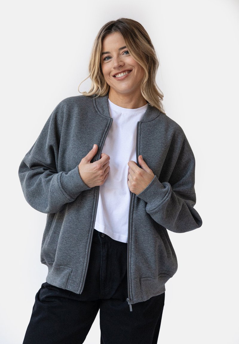 Szary polarowy sweatshirt z zamkiem błyskawicznym, okrągłym dekoltem, ściągaczami na rękawach i bocznymi kieszeniami, połączony z białą koszulką i czarnymi spodniami.