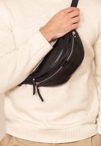 Sac bandoulière en cuir noir avec double fermeture éclair, forme arrondie et sangle réglable, porté contre un pull de couleur claire.