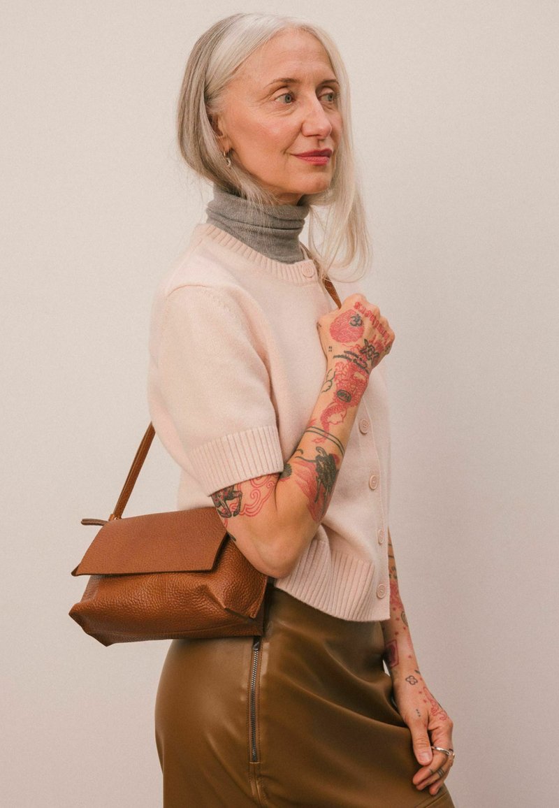 Oudere vrouw met lang grijs haar, kleurrijke tatoeages op armen en handen, draagt een beige korte mouw trui, een bruine leren rok en heeft een bruine handtas bij zich.