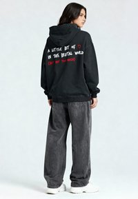 Felpa nera oversize con testo bianco e rosso sul retro. Abbinata a jeans grigi larghi e sneaker bianche. Tessuto morbido, vestibilità casual.