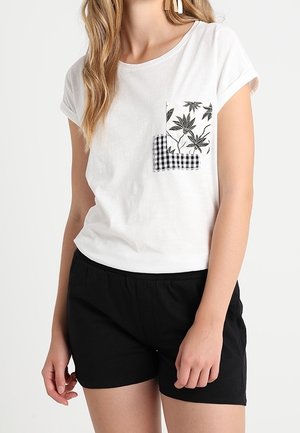 Kvinde iført en hvid t-shirt med korte ærmer med blomster- og ternet lomme og sorte shorts, stående med afslappet kropsholdning.