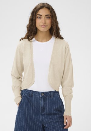 Žena s dugom kovrčavom kosom, nosi bež kratki kardigan preko bijele majice i plave hlače visokog struka sa uskim prugama, ruke u džepovima.