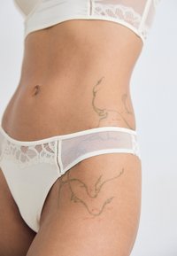 Biały bikini z koronkowym wykończeniem i przezroczystymi panelami siateczkowymi, z niskim stanem i skomplikowanym tatuażem w kwiaty na udzie.