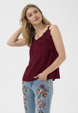 Femme portant un top sans manches en dentelle bordeaux et un jean bleu avec une broderie florale colorée, posant avec une main dans la poche et l'autre touchant ses cheveux.