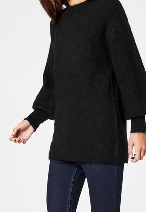 Femme portant un pull noir duveteux ample à manches longues et un jean bleu foncé ajusté devant un fond uni.