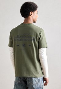 Oliivinvihreä lyhythihainen t-paita, jossa sininen teksti "stockholm ATHLETES CLUB 2015" takana, puuvillamateriaalia, yhdistetty valkoisen pitkähihainen paidan kanssa alla.