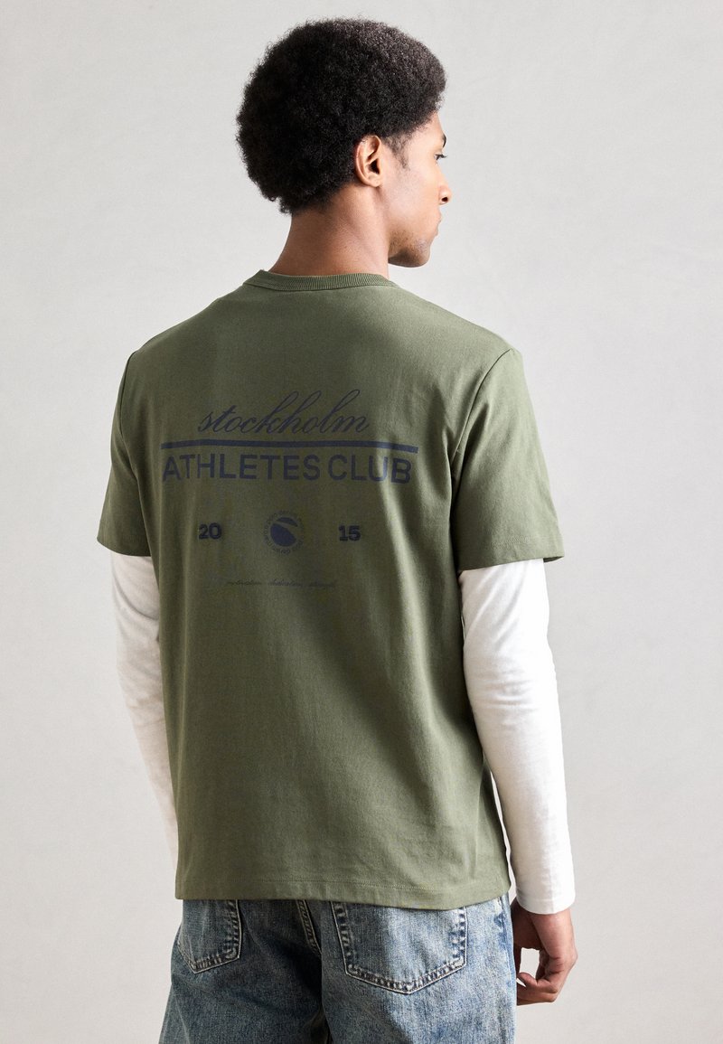 Oliivinvihreä lyhythihainen t-paita, jossa sininen teksti "stockholm ATHLETES CLUB 2015" takana, puuvillamateriaalia, yhdistetty valkoisen pitkähihainen paidan kanssa alla.