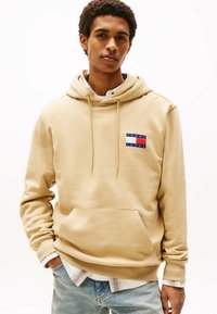 Beige hoodie lavet af blødt materiale, med en frontlomme, snoretræk i huen og et lille flerfarvet logo på brystet. Laget over en hvid skjorte.