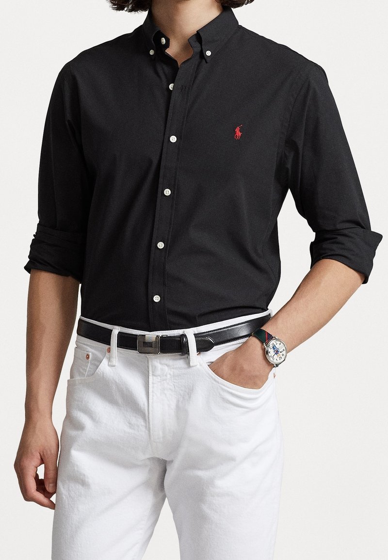 Camicia nera con bottoni e maniche lunghe, dotata di un logo rosso sul lato sinistro del petto, abbinata a pantaloni bianchi e una cintura nera.