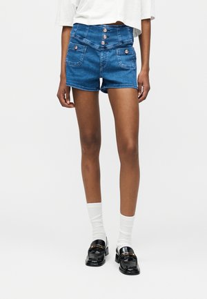 Nainen pukeutuneena korkeavyötäröisiin sinisiin farkkushortseihin, joissa edessä napit, valkoiset sukat, mustat loafersit ja valkoinen lyhyt toppi, seisoo valkoista taustaa vasten.