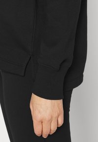 Svart sweatshirt med ribbat mudd och sidoflikar, tillverkad av mjukt tyg. Närbild av ärmen och handen som vilar på svarta leggings.