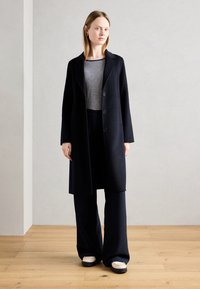 Manteau en laine noir avec de grands boutons, porté sur un haut rayé et un pantalon évasé. Le mannequin se tient sur un sol en bois devant un mur gris.