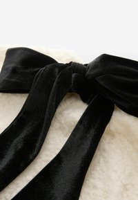 Un lazo de terciopelo negro con colas alargadas, colocado sobre un fondo de tela crema texturizada. Textura suave y lisa con un acabado mate.