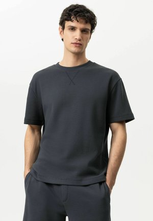 Junger Mann mit lockigem Haar, der ein dunkles, strukturiertes Kurzarmhemd und passende Hosen trägt, steht mit den Händen in den Taschen vor einem weißen Hintergrund.