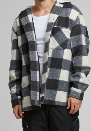 Person trägt eine schwarz-weiß-grau karierte Jacke über einem weißen, gerippten Hemd mit schwarzen Hosen.