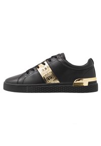 Zapatilla baja negra con cordones negros, suela texturizada y banda decorativa dorada con acento en el talón.