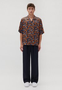 Camicia a maniche corte marrone con stampa leopardata blu e nera, abbinata a pantaloni larghi blu navy e sneakers bianche. Tessuto liscio, design casual.