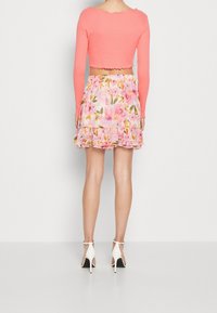 Korallfärgad långärmad kort topp med vågiga kanter, ihop med en rosa blommig volangkjol. Vita högklackade sandaler kompletterar outfiten.