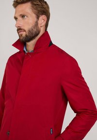 Giacca esterna rossa con colletto alto, caratterizzata da una texture liscia e raffinata. Tra i dettagli degni di nota ci sono un accento a contrasto in tinta blu navy e una tasca con zip.