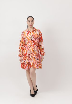 Frau mit langen dunklen Haaren in einem bunten Kleid mit orange-, rosa- und weißen Mustern und schwarzen High Heels, die vor einem schlichten weißen Hintergrund steht.