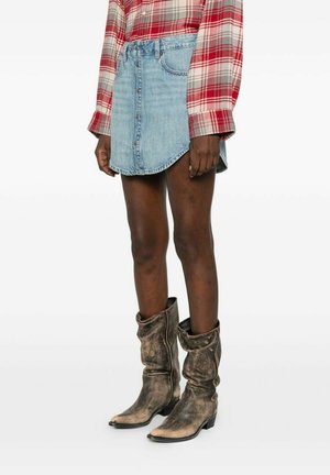 Person trägt ein rot-grau kariertes Hemd, einen hellblauen Jeansrock mit Knöpfen vorne und abgetragene braune Leder-Cowboystiefel.