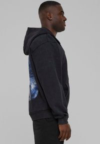 Sort sweatshirt med hætte, med et blåt grafisk design på siden, ribbede manchetter og en kenguru-lomme. Blød tekstur hele vejen igennem.