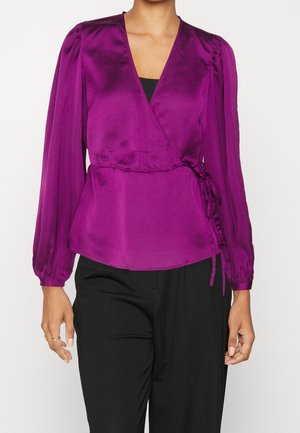Woman wearing a long-sleeve magenta satin wrap blouse tied at the side, paired with black pants, standing against a plain background.