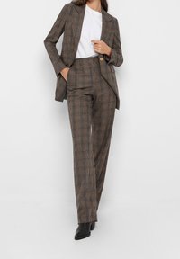 Costume à carreaux marron avec un blazer ajusté et un pantalon à jambes larges. Le tissu a une finition texturée. Associé à une chemise blanche et des bottines noires.