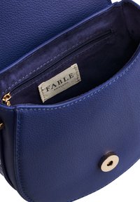 Mala tamnoplava kožna torbica s preklopom na zlatnu tipku, unutarnjim džepom na zatvarač i tkanicom s natpisom "FABLE England."