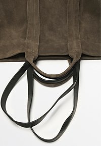 Sac fourre-tout en daim marron avec des poignées en cuir noir lisse, présentant des coutures surpiquées et un design simple et ouvert.