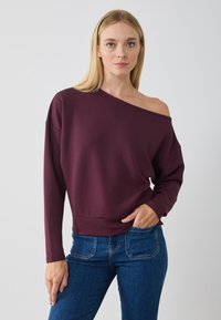 Sudadera burdeos de hombros descubiertos con mangas largas y un ajuste relajado, combinada con jeans azul oscuro, con una textura suave.