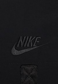 Černé logo Nike s vyšívaným motivem, texturovaná tkanina, obdélníková záplata se šitým detailem a hladké pozadí.