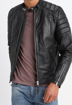 Mężczyzna w czarnej skórzanej kurtce motocyklowej z wieloma kieszeniami na zamek, pod spodem brązowa koszula oraz niebieskie dżinsy.