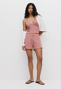 Ljust rosa ribbat stickad linne med v-ringning, parat med matchande shorts och svarta flip-flops; vit jacka hängande över ena axeln.