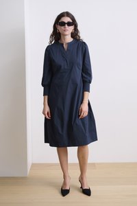 Tumesinine puuvillane kleit, millel on lõhikiga krae, narmastega varrukad ja lõdva lõige. Kombineeritud mustade teravaotsaga kingade ja nelinurksete prillidega.