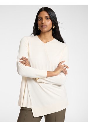 Frau mit langem dunklem Haar, die goldene Creolen trägt, ein cremefarbenes asymmetrisches Langarmshirt und olivgrüne Hosen, mit verschränkten Armen und neutralem Gesichtsausdruck.