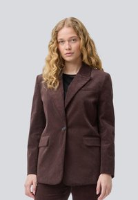 Giovane donna con lunghi capelli ricci biondi che indossa un blazer di velluto a coste marrone scuro e pantaloni abbinati su uno sfondo semplice.