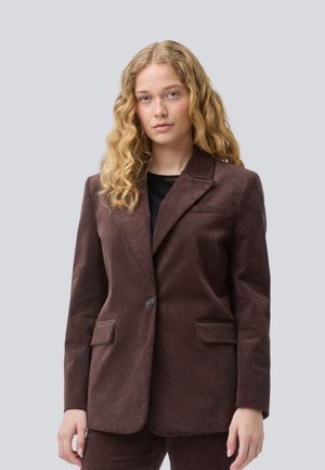 Junge Frau mit langen lockigen blonden Haaren, die ein dunkelbraunes Cord-Blazer und passende Hose vor einem schlichten Hintergrund trägt.