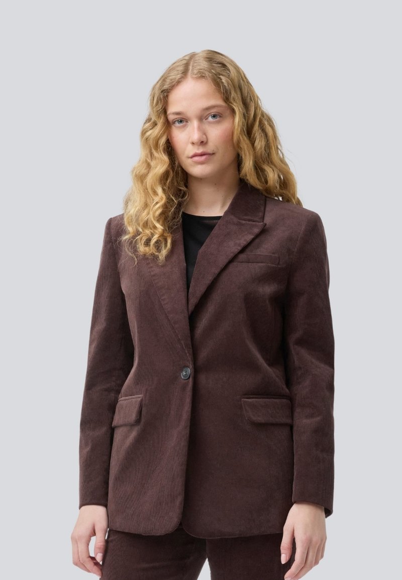 Giovane donna con lunghi capelli ricci biondi che indossa un blazer di velluto a coste marrone scuro e pantaloni abbinati su uno sfondo semplice.