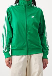 Grön zip-up träningsoveralljacka i mjukt tyg. Har vita tre-rands detaljer på ärmarna och en liten logotyp på bröstet.