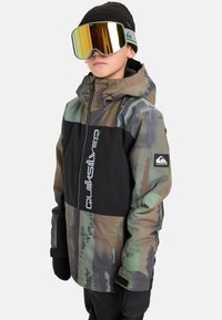 Kamufliažinė striukė su juodu juostu, su užtrauktuku priekyje, reguliuojama kapisonu ir Quiksilver logotipu. Derinama su auksinėmis veidrodinėmis akinių.