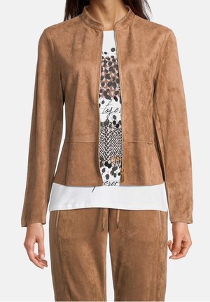 Mujer con una chaqueta de ante marrón claro con cremallera, sobre una camiseta blanca con diseño gráfico y pantalones de ante marrón a juego.