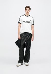 Biely tričko Adidas s čiernymi akcentmi a logom, v kombinácii s čiernymi nohavicami z umelého kože a bielymi teniskami. Čierna šiltovka držaná v ľavej ruke.