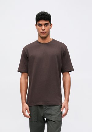 Brun, kortärmad T-shirt i bomull med rund hals och en diskret logodetalj på bröstet. Modellen bär olivgröna byxor.