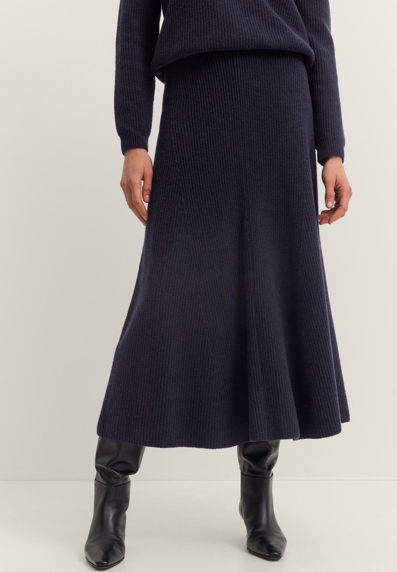 Marineblaue Strickrock mit gerippter Textur, ausgestelltem Design, bis zur Mitte der Wade reichend, kombiniert mit knielangen schwarzen Lederstiefeln.