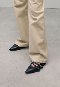 Zwarte puntige schoenen met een enkelriem met metalen oogjes en een gesp, gecombineerd met beige wijde broekspijpen.