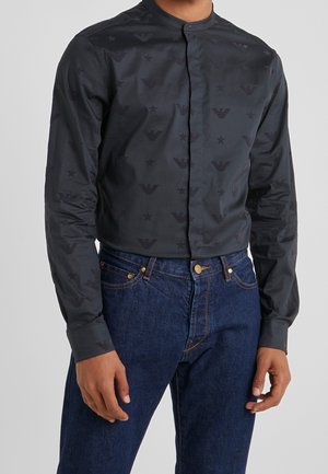 Mężczyzna ubrany w czarną koszulę z kołnierzykiem mandarynkowym z delikatnymi wzorzystymi logo, w połączeniu z ciemnoniebieskimi jeansami, stojący na jednolitym tle.